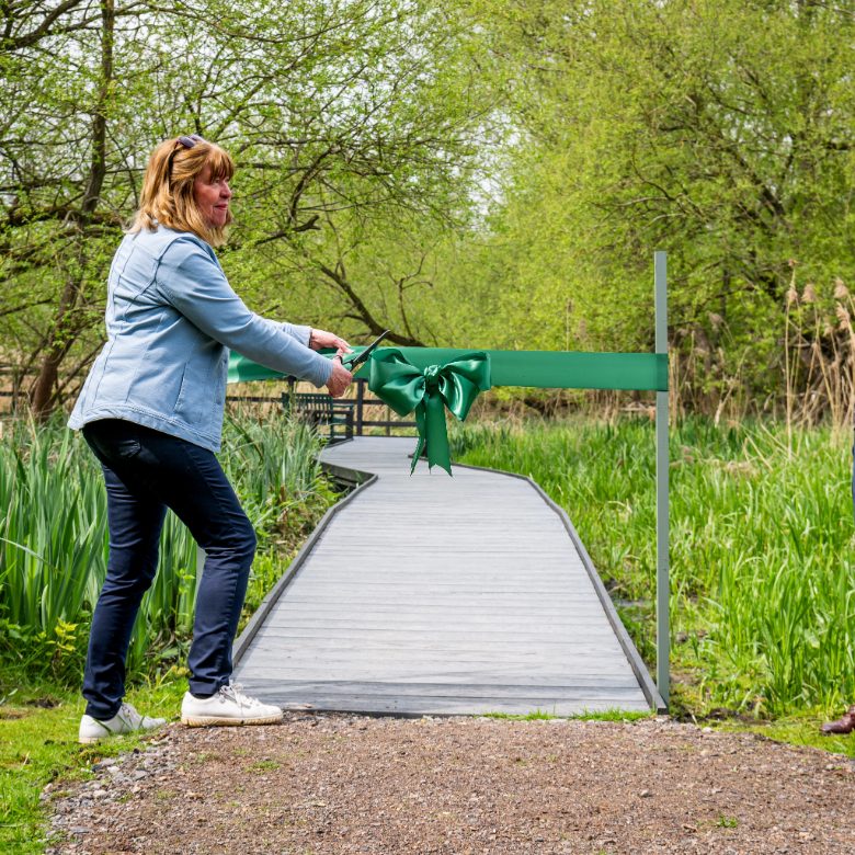 Rebecca Joisce, former Chair and trustee, smiling, cuts a green ribbon to open the new boardwalk to visitors.