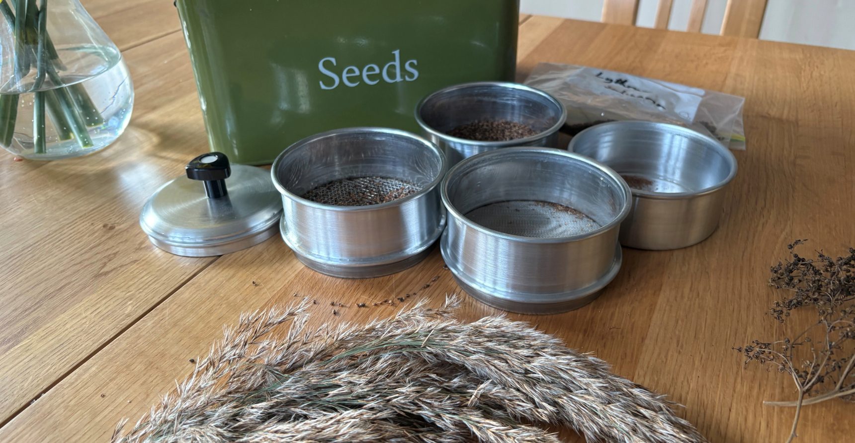 A round metal seed sieve separated into its four sections is placed on a table in front of a metal seed box, and dried seedheads of reeds and other plant material.