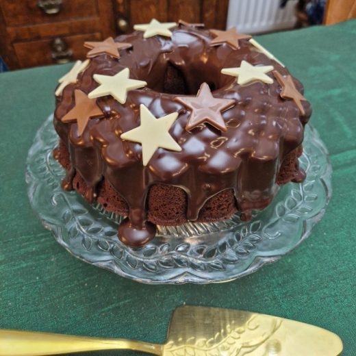 A delicious looking chocolate orange bundt cake sits on a glass cake stand, with a gold cake slice at the ready. The cake is covered with chocolate ganache icing, and white, dark and milk chocolate stars adorn the top.